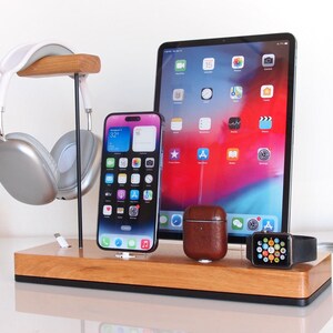 Può includere: Una stazione di ricarica in legno con una base nera, dotata di un supporto per cuffie argentato, un tappetino di ricarica wireless bianco, una custodia in pelle marrone per AirPods, un supporto di ricarica Apple Watch nero e un supporto di ricarica iPhone nero. La stazione mostra un iPhone nero, una custodia in pelle marrone per AirPods e un Apple Watch nero.