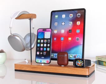 May include: A wooden charging station with a black base, featuring a silver headphone stand, a white wireless charging pad, a brown leather case for AirPods, a black Apple Watch charging stand, and a black iPhone charging stand. The station is displaying a black iPhone, a brown leather AirPods case, and a black Apple Watch.