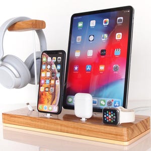 May include: A wooden charging station with a tablet, smartphone, wireless earbuds, and a smartwatch. A pair of gray headphones hangs on a wooden stand. The tablet displays a colorful app screen.