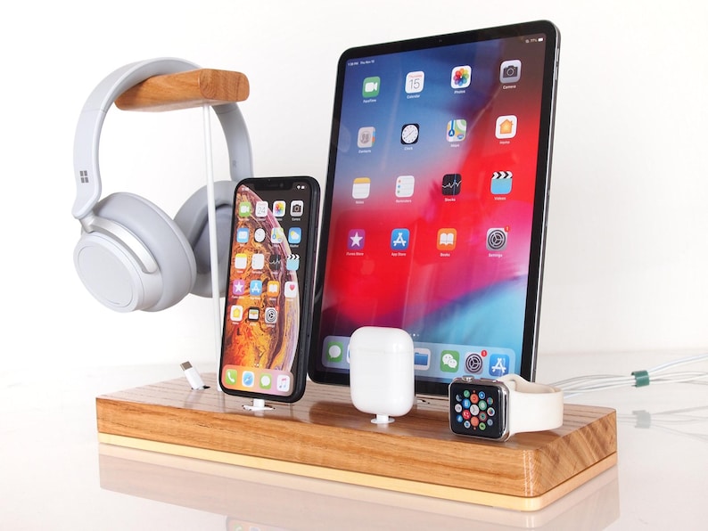 May include: A wooden charging station with a tablet, smartphone, wireless earbuds, and a smartwatch. A pair of gray headphones hangs on a wooden stand. The tablet displays a colorful app screen.