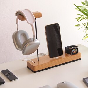 May include: Wooden charging station with a light pink and silver headset, a black smartphone, and a black smartwatch. The station also holds a white charging cable. The desk has two black remotes and a computer mouse.