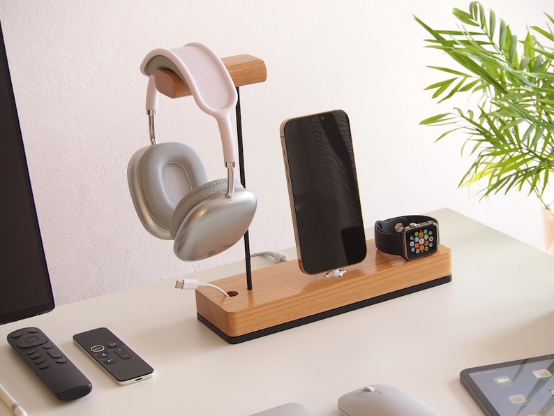 May include: Wooden charging station with a light pink and silver headset, a black smartphone, and a black smartwatch. The station also holds a white charging cable. The desk has two black remotes and a computer mouse.