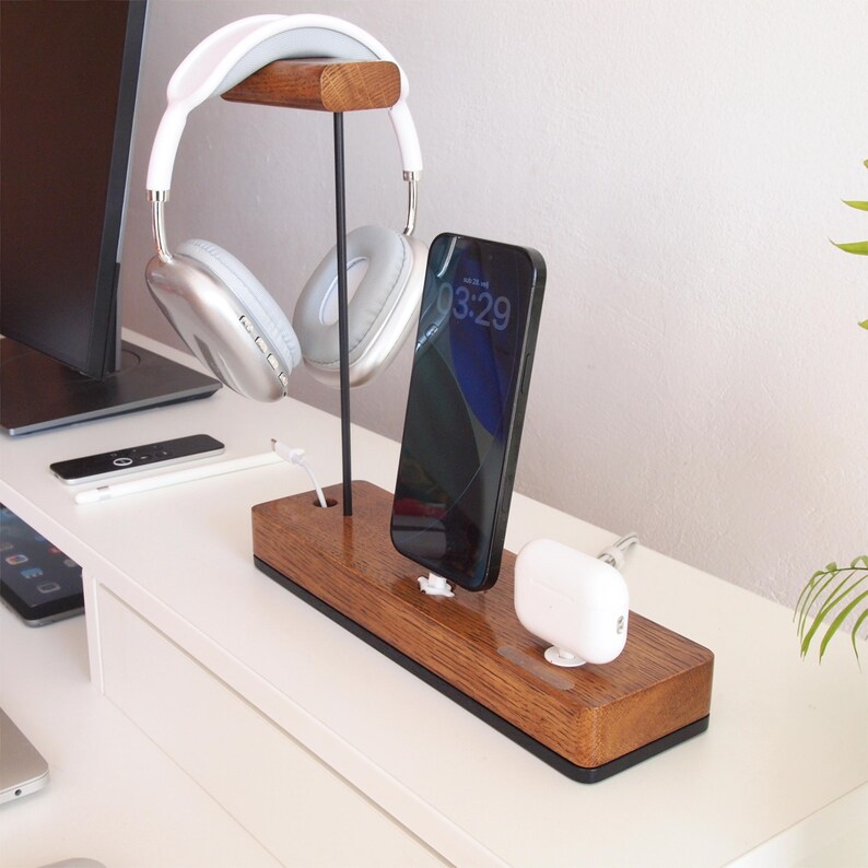 May include: A wooden desk organiser featuring a headphone stand, a smartphone, and a charging station for wireless earbuds. The headphones are white and silver. The smartphone displays the time 03:29.