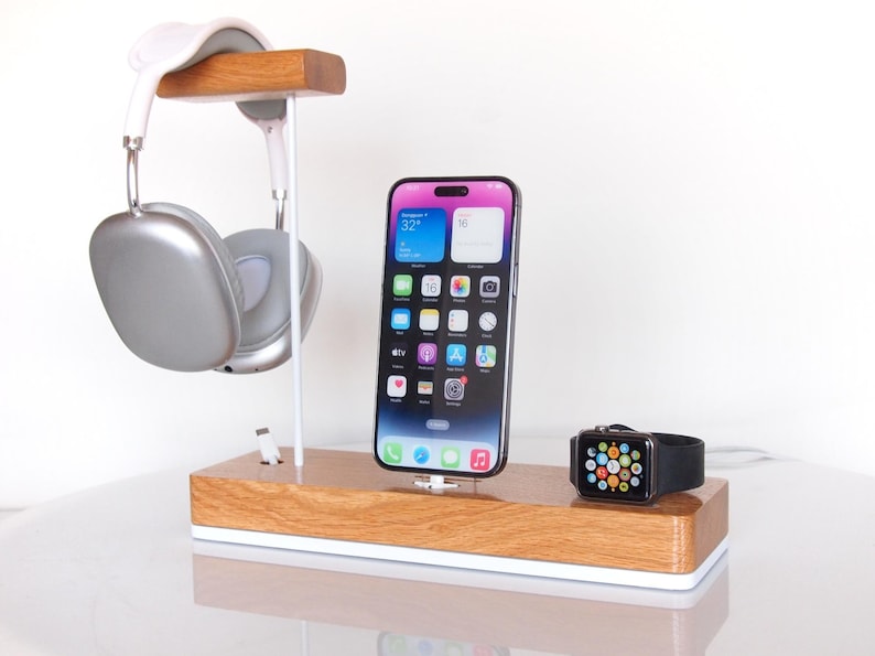 May include: Wooden charging station with a white base, holding a silver headset, a smartphone, and a smartwatch. The phone displays app icons. The headset is hanging from a wooden bar. The smartwatch has a black strap.
