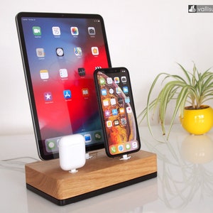 Può includere: Una stazione di ricarica in legno con una base nera, che tiene un iPad, uno smartphone e un set di auricolari wireless. La stazione è su una superficie bianca.