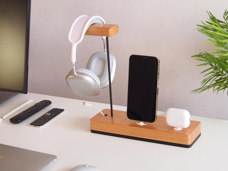 May include: A wooden desk organiser featuring a headphone stand, phone holder, and space for wireless earbuds. The stand holds silver and white headphones, a black smartphone, and white earbuds. A white charging cable is plugged in.