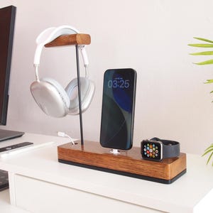 May include: A wooden desk organiser with a headphone stand, phone charger, and watch charger. The headphones are white and silver. The phone screen displays 03:25. The watch has a black strap.