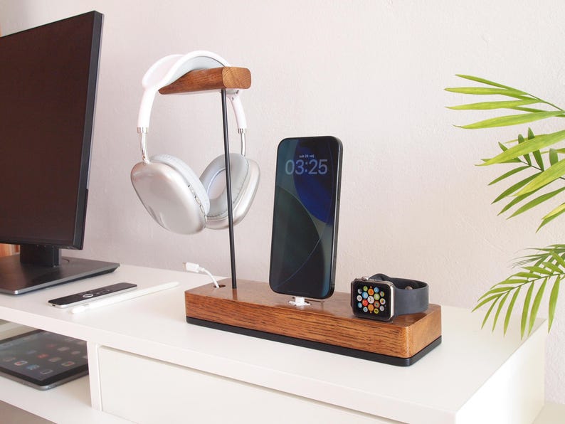May include: A wooden desk organiser with a headphone stand, phone charger, and watch charger. The headphones are white and silver. The phone screen displays 03:25. The watch has a black strap.
