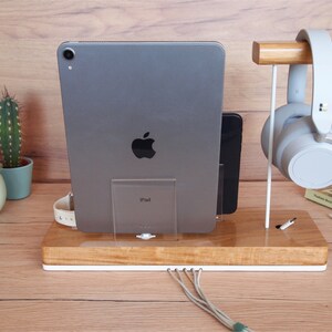 May include: A wooden desk organizer with a white base, holding a gray iPad, a black smartphone, and a white smartwatch. A pair of gray headphones hangs on a wooden stand. Cables extend from the base. A cactus in a pot is visible in the background.