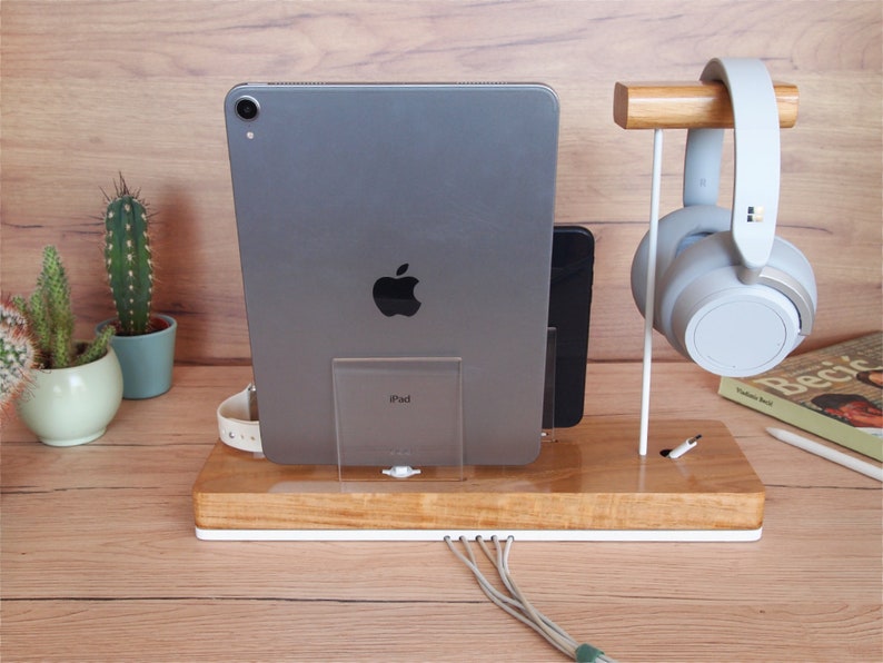 May include: A wooden desk organizer with a white base, holding a gray iPad, a black smartphone, and a white smartwatch. A pair of gray headphones hangs on a wooden stand. Cables extend from the base. A cactus in a pot is visible in the background.