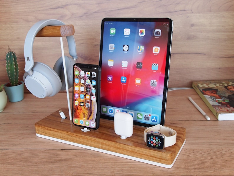 May include: A wooden charging station with a white base holds a tablet, smartphone, smartwatch, and wireless earbuds. A pair of gray headphones rests on a wooden stand. A cactus in a small pot is visible.