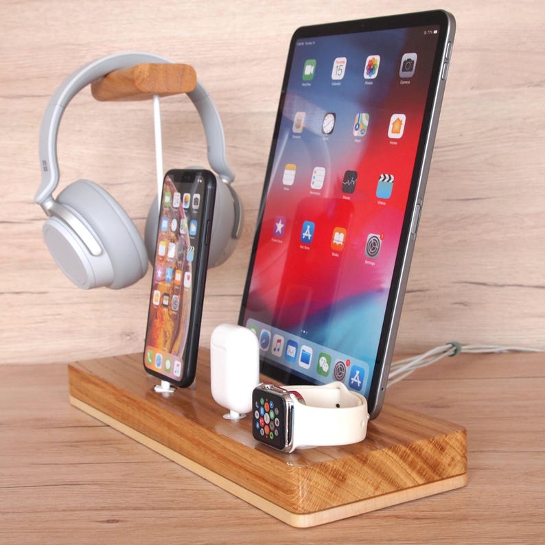 May include: Wooden charging station with a tablet, smartphone, smartwatch, wireless earbuds, and headphones. The tablet displays app icons. The smartwatch has a white band. The headphones are gray.