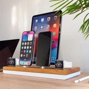 May include: A wooden charging station with a white base holds a tablet, two smartphones, and two smartwatches. A laptop and accessories are on the wooden desk. The devices are charging. The background is a white wall with a green plant.