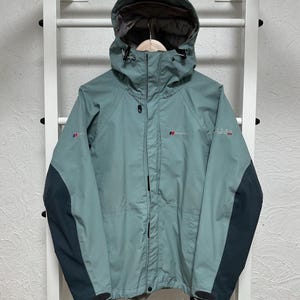 Może przedstawiać: Teal i ciemnoszara kurtka z kapturem z nazwą marki "Berghaus" na piersi. Kurtka ma zamek błyskawiczny na całej długości, sznurki u dołu i ciemnoszare panele na rękawach. Kurtka wisi na białym stojaku.