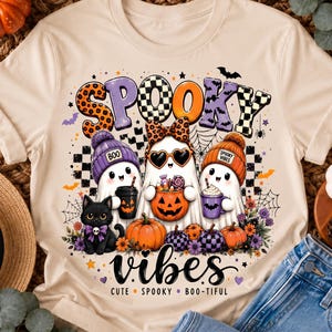 Może przedstawiać: Kremowy t-shirt z grafiką przedstawiającą trzy duszki w czapkach, czarnego kota i dynie. Napis głosi SPOOKY VIBES, CUTE, SPOOKY, BOO-TIFUL. Duszki trzymają kubek kawy, wiaderko w kształcie dyni ze słodyczami oraz filiżankę.