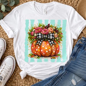 May include: A white short-sleeve t-shirt featuring a central graphic of an orange pumpkin with white polka dots, topped with a large black and white buffalo plaid bow and a vibrant arrangement of pink, orange, and yellow flowers with green leaves.