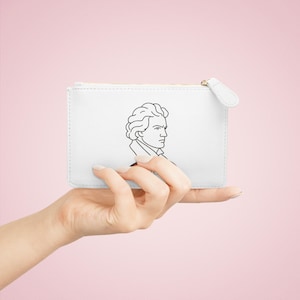 Könnte beinhalten: Weiße Reißverschlusstasche mit einer schwarzen Strichzeichnung eines Männerprofils. Die Tasche wird in einer Hand vor einem rosa Hintergrund gehalten. Die Tasche hat einen weißen Reißverschluss.