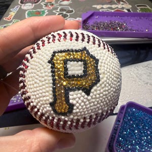 May include: A white baseball with red stitching and a large, gold and black letter "P" made of sparkling rhinestones. The baseball is covered in small, white rhinestones.