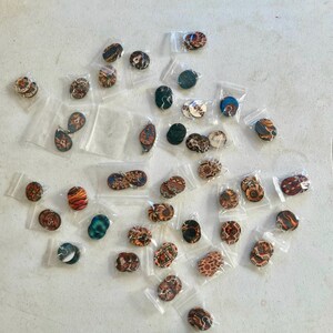 May include: A collection of small, patterned earrings and pendants in clear plastic bags. The earrings and pendants feature various designs, including swirls, geometric patterns, and abstract shapes. The color palette includes shades of brown, orange, blue, and green.