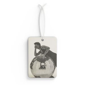 May include: Rectangular air freshener featuring a black and white image of a person leaning on a disco ball. The air freshener has a white string for hanging.