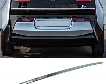 May include: Rear view of a black and silver BMW i3 electric car. The car has a silver trim piece and the model name "i3" is visible. The taillights are illuminated with red and white lights. The BMW logo is centered above the license plate area.