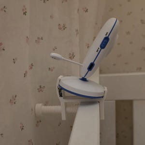 Könnte beinhalten: Ein weiß-blauer Babyphone-Monitor, der an einer weißen Kinderbett-Schiene befestigt ist. Der Monitor hat ein einstellbares Mikrofon und einen Ein-/Ausschalter. Der Markenname "Suze" ist auf der Klemme sichtbar.