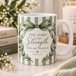 May include: A white ceramic mug featuring a sage green vertical striped pattern. The front displays a sage green ribbon bow, floral accents, and the text: THE LORD IS MY STRENGTH AND MY SHIELD, PSALM 28:7, with a small heart icon centered below.