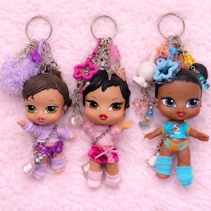 Puede incluir: Tres llaveros de muñecas Bratz. Cada muñeca tiene ojos grandes, labios carnosos y viste un atuendo diferente. Los llaveros tienen varios dijes y cuentas. Las muñecas están sobre una superficie rosa y difusa.