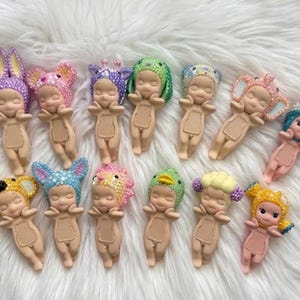 May include: Thirteen small figurines with light skin tones, each wearing a unique headpiece covered in colorful rhinestones. The figures have closed eyes, small smiles, and a rectangular indentation on their chests, arranged in two rows.