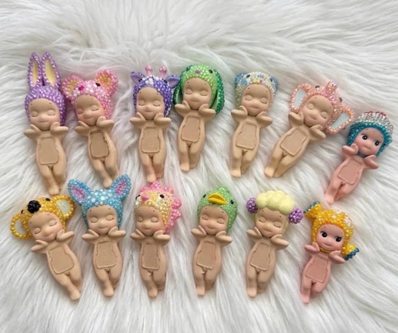 May include: Thirteen small figurines with light skin tones, each wearing a unique headpiece covered in colorful rhinestones. The figures have closed eyes, small smiles, and a rectangular indentation on their chests, arranged in two rows.