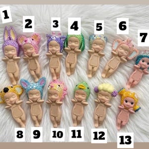May include: Thirteen small, light-skinned baby figurines arranged in two rows, each wearing a unique, colorful, textured headpiece. The figures are numbered one through thirteen, featuring various animal and floral-inspired designs with beads.