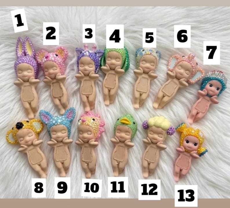 May include: Thirteen small, light-skinned baby figurines arranged in two rows, each wearing a unique, colorful, textured headpiece. The figures are numbered one through thirteen, featuring various animal and floral-inspired designs with beads.