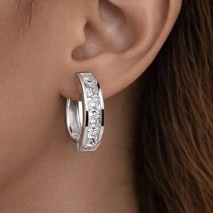 May include: A silver hoop earring with a row of square, clear gemstones. The earring is worn on a person's ear, with the skin and hair visible in the background. The earring is a classic jewelry piece.