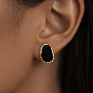 Puede incluir: Primer plano de una oreja con un pendiente de botón dorado. El pendiente tiene un centro negro de forma irregular y un borde dorado. El pendiente es de tipo botón.