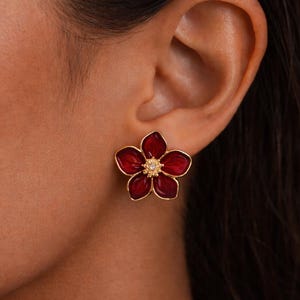 Peut inclure: Gros plan d'une boucle d'oreille à clou en forme de fleur rouge avec un contour doré et un petit centre scintillant. La boucle d'oreille est portée sur une oreille sur fond sombre.