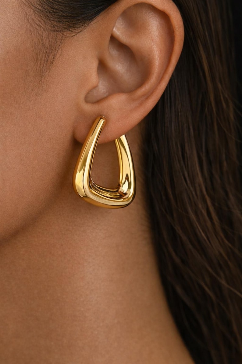 K&ouml;nnte beinhalten: Nahaufnahme eines goldfarbenen, tropfenf&ouml;rmigen Ohrrings. Der Ohrring ist aus dickem, massivem Metall mit glatter, reflektierender Oberfl&auml;che. Der Ohrring wird am Ohr einer Person getragen.