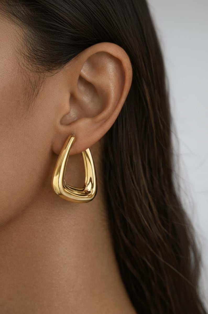 K&ouml;nnte beinhalten: Nahaufnahme eines goldfarbenen, tropfenf&ouml;rmigen Ohrrings. Der Ohrring wird am Ohr einer Person mit dunkelbraunem Haar getragen. Der Ohrring hat eine glatte, polierte Oberfl&auml;che und ein hohles Innere.