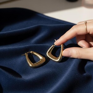 K&ouml;nnte beinhalten: Ein Paar goldfarbene, geometrische Creolen. Die Ohrringe haben ein einzigartiges, eckiges Design mit einer abgerundeten Innenkante und einer flachen Au&szlig;enkante, was eine moderne, minimalistische &Auml;sthetik schafft. Die Ohrringe werden auf einem dunkelblauen, seidigen Stoff pr&auml;sentiert.