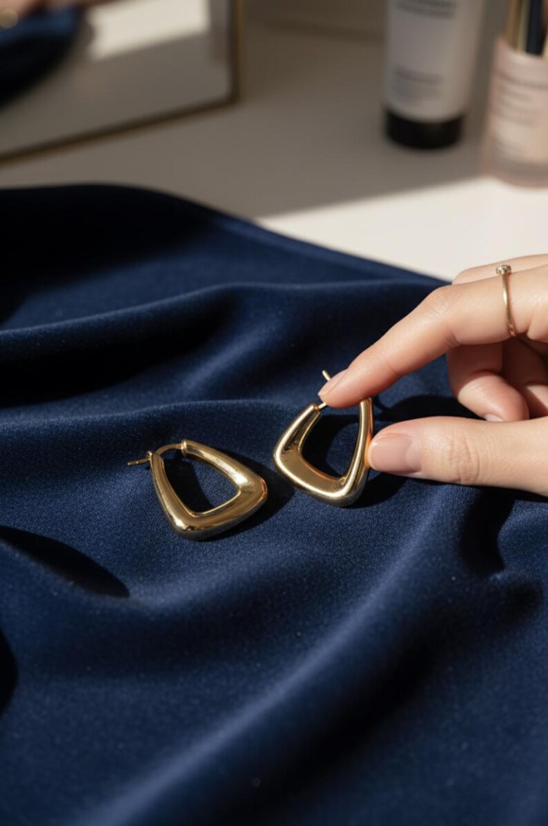 K&ouml;nnte beinhalten: Ein Paar goldfarbene, geometrische Creolen. Die Ohrringe haben ein einzigartiges, eckiges Design mit einer abgerundeten Innenkante und einer flachen Au&szlig;enkante, was eine moderne, minimalistische &Auml;sthetik schafft. Die Ohrringe werden auf einem dunkelblauen, seidigen Stoff pr&auml;sentiert.