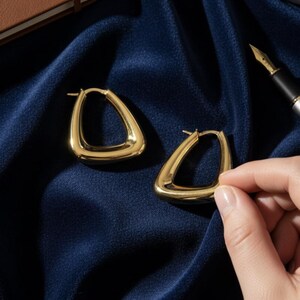K&ouml;nnte beinhalten: Ein Paar goldfarbene, dreieckige Creolen. Die Ohrringe werden auf einem dunkelblauen, seidigen Stoff pr&auml;sentiert. Eine Hand h&auml;lt einen der Ohrringe. Ein braunes Notizbuch, ein Stift und eine Kaffeetasse befinden sich im Hintergrund.