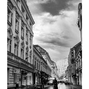 Könnte beinhalten: Schwarz-Weiß-Fotografie einer europäischen Stadtstraße. Gebäude säumen beide Seiten der Straße, mit einer Person, die im Vordergrund Fahrrad fährt. Der Himmel ist bewölkt.