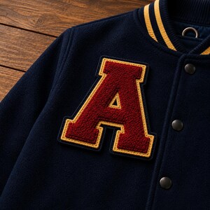 May include: A navy blue varsity jacket featuring a large, textured maroon letter A patch with a yellow border. The jacket includes a ribbed collar with yellow stripes, dark metal snap buttons, and a soft, fleece-like fabric construction.