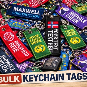 Może przedstawiać: Kolekcja kolorowych breloków z różnymi logo i tekstami, w tym "Maxwell Construction", "Midvale Cheer" i "Your Logo Here". Etykiety są w kolorach czerwonym, niebieskim, zielonym i fioletowym, ze srebrnymi kółkami. Na zdjęciu znajduje się również pilot do samochodu.