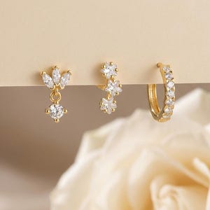 May include: Three pairs of gold-colored earrings. One pair features a dangling, teardrop-shaped design with a round, clear stone. Another pair has a floral design with clear stones. The third pair is a hoop earring with clear stones.