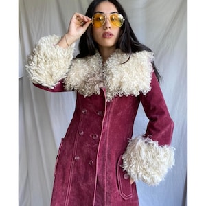 May include: A deep burgundy suede coat featuring a double-breasted front with matching buttons, long sleeves, and a large, fluffy cream-colored curly faux fur collar and matching oversized cuffs on the sleeves, creating a textured contrast.