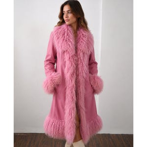 Könnte beinhalten: Ein langer, leuchtend pinker Mantel mit glatter, matter Oberfläche und üppigem, zotteligem pinkem Kunstfellbesatz an den breiten Revers, den Ärmelbündchen und am unteren Saum, was für ein strukturiertes und geschichtetes Erscheinungsbild sorgt.