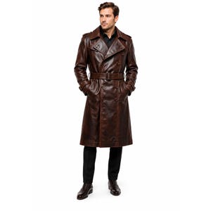May include: A dark brown leather trench coat featuring a double-breasted button front, a matching waist belt with a buckle, wide lapels, shoulder epaulets, and long sleeves with buckled cuffs, worn over a black shirt and black trousers.