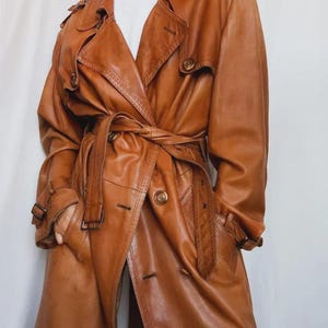 Puede incluir: Un abrigo trench de cuero marrón con cinturón en la cintura, botones y charreteras. El abrigo tiene un diseño clásico con cuello y bolsillos. El abrigo es de color marrón medio.