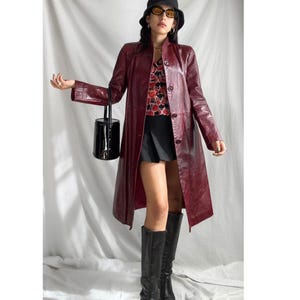 Peut inclure: Un long manteau en cuir rouge foncé avec une fermeture boutonnée, porté sur un haut à motifs et une mini-jupe noire. La tenue comprend un bob noir, des lunettes de soleil teintées de jaune, un sac à main cylindrique noir et des bottes hautes.