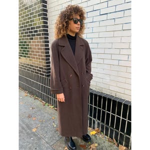 Puede incluir: Un abrigo largo marrón de doble botonadura con un cuello de solapa clásico. El abrigo es hasta la rodilla y se lleva con zapatos negros. El abrigo está hecho de un material similar a la lana y tiene un corte entallado.